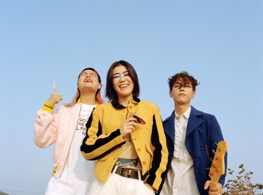 The three members of the Korean indie band SE SO NEON. The picture is taken from the hip up; two of them are laughing and the other one is holding the neck of an electric guitar.