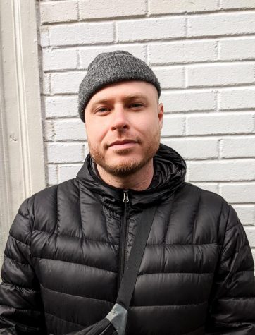 A portrait of Misha Tyutyunik against a beige brick wall. He is wearing a black puffer jacket, a fanny pack across his chest and a gray beanie.