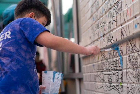 A young boy adds blue paint to a public mural. He is wearing a blue t-shirt and holds a large plastic container in his hand.