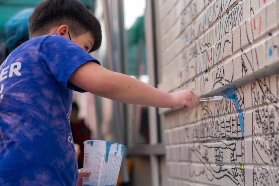 A young boy adds blue paint to a public mural. He is wearing a blue t-shirt and holds a large plastic container in his hand.