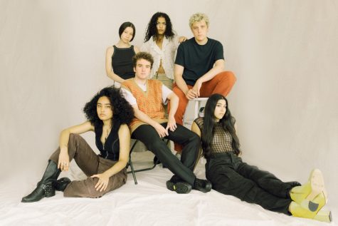 A group studio portrait of six singers from the music collective MICHELLE in front of a white seamless backdrop. Two members sit on the ground, while the other four sit on stools of alternating height.