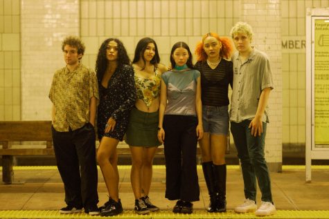 A group portrait of six singers from the music collective MICHELLE. They are standing on a subway platform in front of a brown bench.