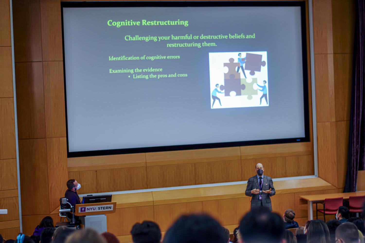 A large lecture hall full of students. On the bottom right, a professor addresses the class. On the bottom left, a course assistant sits behind a podium. Above them, a slide presentation is displayed on a large screen with information about cognitive restructuring.