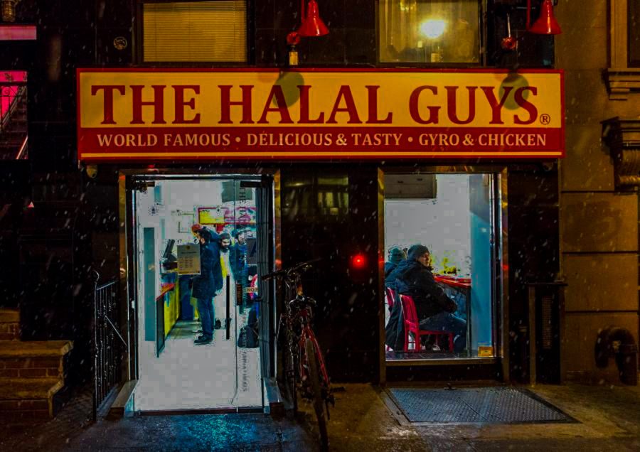 The exterior façade of the brown building with the red, yellow and white signage of The Halal Guys storefront.