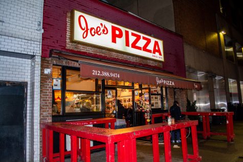 The exterior façade of a red brick building with a Joe's Pizza sign and patio cover while a customer eats at the outside table.