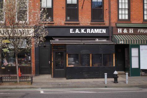 The all black facade of E.A.K Ramen. Above the door, the restaurant’s name hangs in bold white letters. To the left of the entrance, a white apple blossom tree is in full bloom.