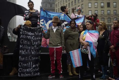 A group of protesters in Foley Square at the Civic Center. On the right, a black banner with the names of trans people of color painted in white. In the center behind a row of people is a banner with blue, white and pink details. On the right, a lady stands with a trans flag tied around her shoulders like a cape.