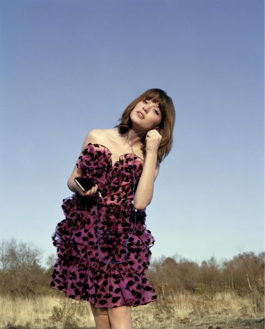 Maisie Peters in a short pink dress with black polka dots, holding a cell phone in her right hand and wired headphones in her left.