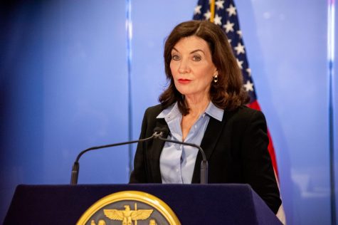 Gov. Kathy Hochul speaks at a podium during a press conference. She stands in front of an American flag and a royal blue background. She is wearing a black blazer with a pale blue collared shirt.