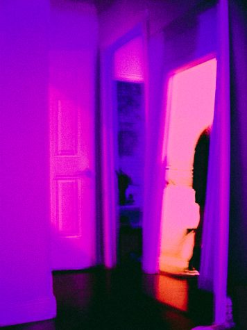 A hallway in pink and purple light with a mirror reflecting back into a bedroom.