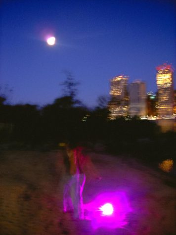 A blurry beach and city skyline with the moon overhead. Someone in motion on the sand next to purple light.