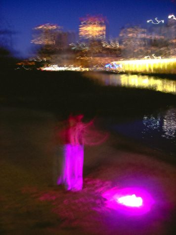 A blurry beach and city skyline with the moon overhead. Someone in motion on the sand next to purple light.