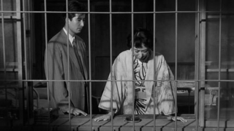 A black-and-white picture of actress Yumeji Tsukioka behind metal bars, looking down at her hands on a table. To her left, her husband is standing and looking at her.