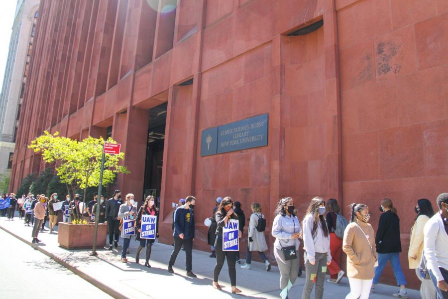 Students march in line on the white concrete sidewalk on the south side of West Fourth Street in front of the red Bobst library building passing by a small green tree and a red street sign. Some of the students hold posters in support of the Graduate Student Organizing Committee.