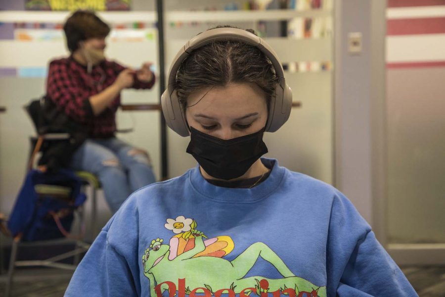 A student seated indoors, wearing a black mask, tan headphones, and a blue sweater with a reclining green frog.