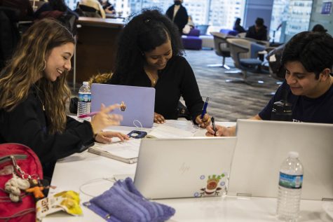 A group of unmasked, smiling students sit at a table in the Kimmel Center for University Life, pens in hand, writing. Their laptops are open.