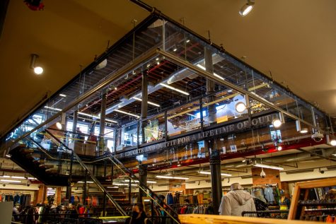 The interior of the REI SoHo Flagship store. The photograph is taken from a low angle, looking from one level up a staircase to a higher level. Clothing racks can be seen throughout the store in the background, and there is a mannequin wearing a hoodie in the foreground.