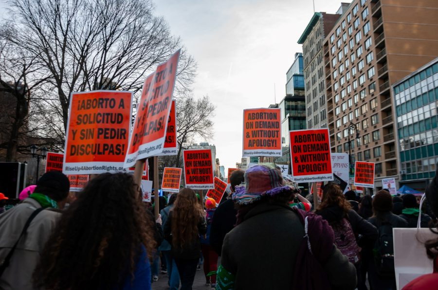 A picture of multiple women at a rally walking on the sidewalk on their way to Washington Square Park, holding posters with an orange background. The posters say in black bold letters: “Abortion on Demand & Without Apology!”