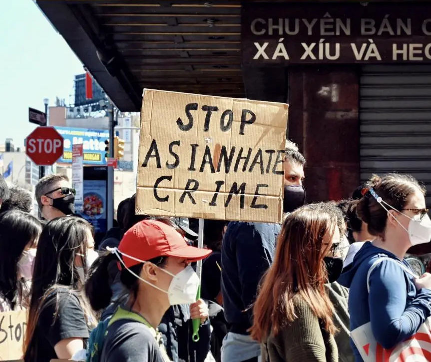 A group of protesters march through Chinatown. In the middle of the frame, one protester holds a cardboard sign with the words “Stop Asian Hate Crime” painted in thick black letters.