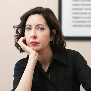 Picture of Sarah Manguso. She has shoulder-length brown curly hair, red lipstick and is looking directly at the camera with her hand on her chin.