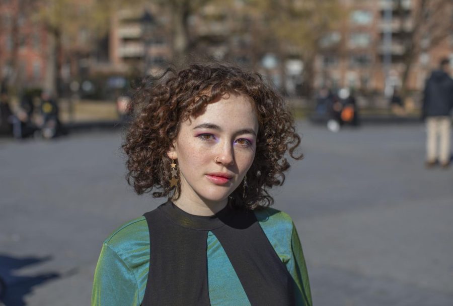 A portrait of Stella Wunder in Washington Square Park. They are wearing an iridescent turquoise shirt with a black vest on top. She is also wearing pink eyeliner and gold star earrings.