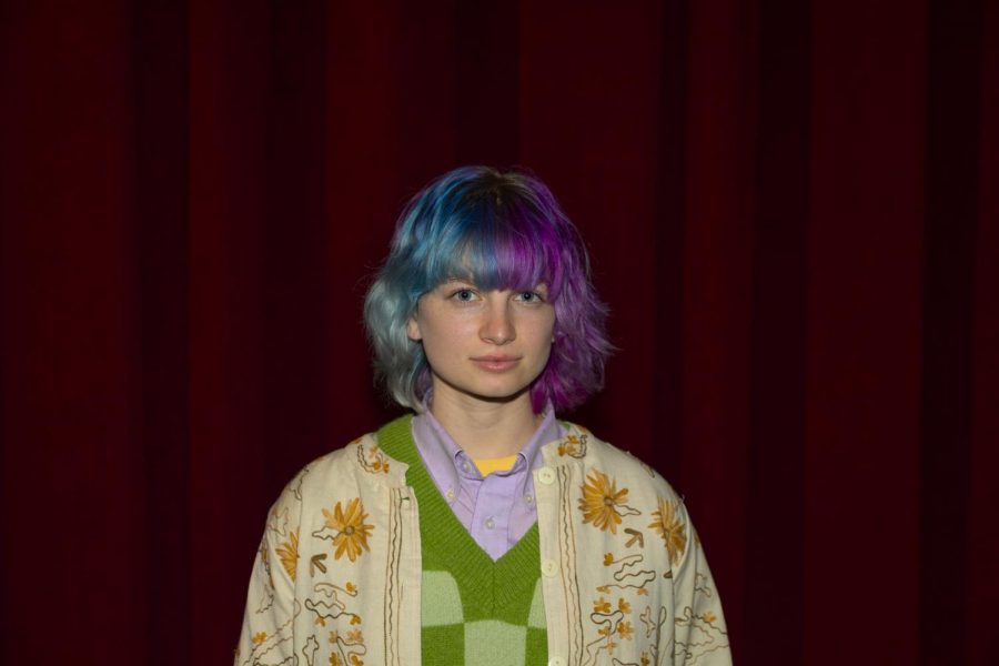 A portrait of Casey Hall-Landers posing in front of a red velvet curtain. They are wearing a beige jacket with orange embroidered flowers, a green checkered vest, and a purple collared shirt. Additionally, they have split dyed hair with blue on the left and pink on the right.