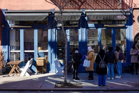 The blue facade of the While We Were Young restaurant. Tables and chairs are placed outdoors on the left of the entrance. On the right, people wait in line to enter.