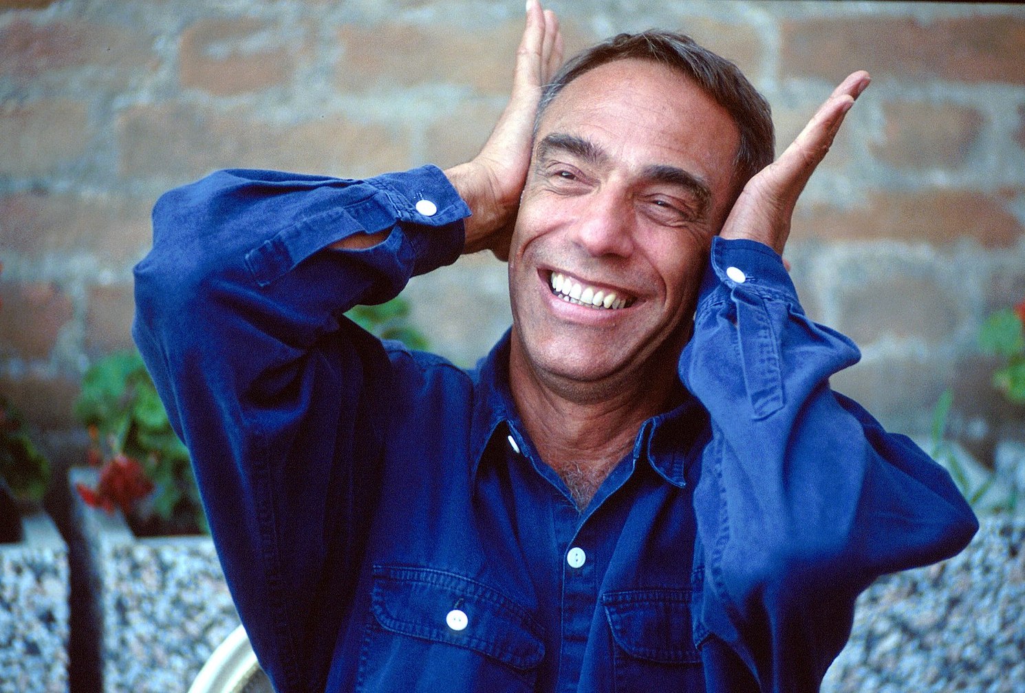 Film director Derek Jarman pictured sitting outdoors at the Venice Film Festival in 1991, wearing a blue button-up shirt. He is smiling and holding up the palms of his hands against the sides of his head.