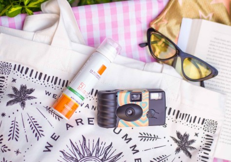 A white tote bag with black decorations, sunscreen, a disposable camera, black glasses with yellow lenses, and an open book on top of a pink checkered picnic blanket.