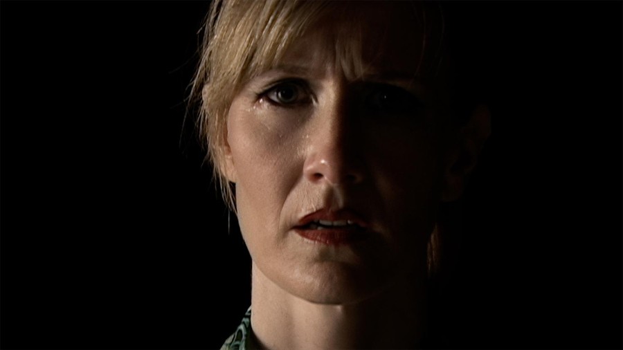 A photo of a womans face staring straight ahead. She is wearing red lipstick and other makeup, and appears to be frightened.