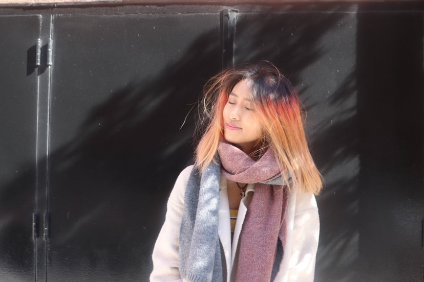 Aria+Young+stands+with+her+eyes+closed+against+black+doors.+She+is+wearing+a+gray+jacket+and+a+blue-and-purple+scarf.+Her+hair+is+a+gradient+of+black%2C+red%2C+orange+and+yellow.
