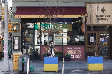 A front view of the Ukrainian restaurant Veselka. The restaurant has a small dark red and yellow awning with its name printed on it. In front of the restaurant there are trees without branches in pots painted with the yellow-and-blue Ukrainian flag.