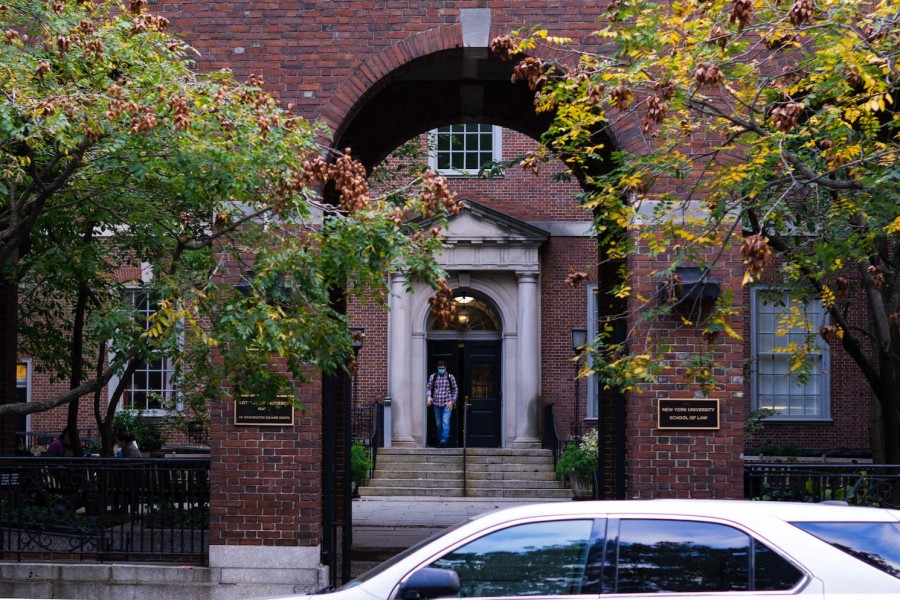 A photo of the outside of the NYU Law School building. A man wearing a mask is seen walking outside the front door into the courtyard.