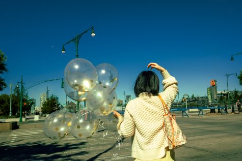 The back of a woman holding balloons as she walks down the street. The balloons are transparent with golden glitter inside. The woman has short black hair and is wearing a white sweater.