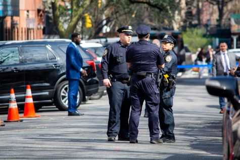 Three NYPD officers in their dark blue uniforms congregating in the middle of the road appear to be discussing something. The officers are flanked by legal personnel dressed in suits on each side.