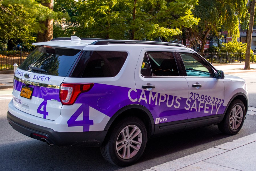 An NYU Campus Safety vehicle in front of trees. The vehicle is a white car with purple decals and the text “Campus Safety 212-998-2222” on top.
