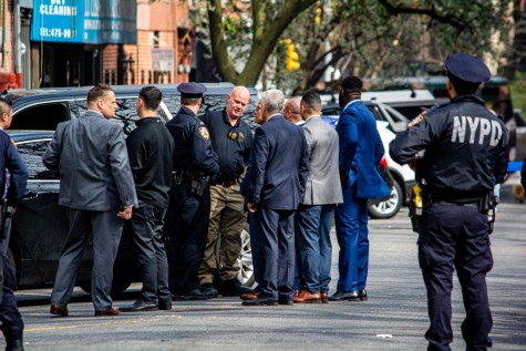 A group of police officers stand grouped on the street. Some wear uniforms while others wear suits. An officer in uniform to the right of the image overlooks the group, with the text “NYPD” visible on the back of the uniform.