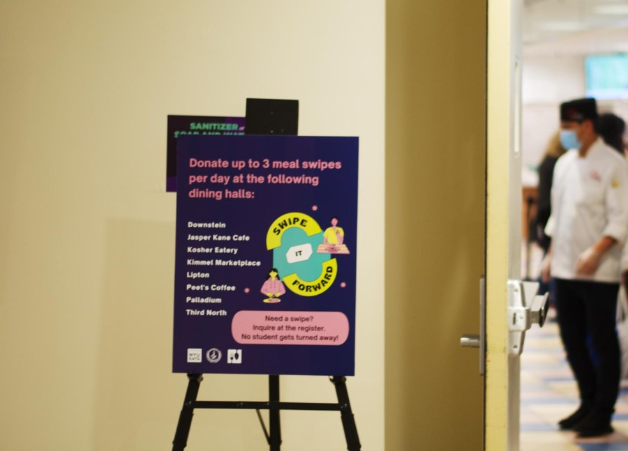 On an easel, a poster with a dark purple background reads in pink font: “Donate up to 3 meal swipes per day at the following dining halls.” Below in white font, one under the other, it reads: “Downstein, Jasper Kane Cafe, Kosher Eatery, Marketplace at Kimmel, NYU Eats at Lipton, Peet’s Coffee, Palladium, NYU Eats at Third North.” Next to the easel the door of Kimmel Marketplace is open and a dining hall worker is seen in the back.