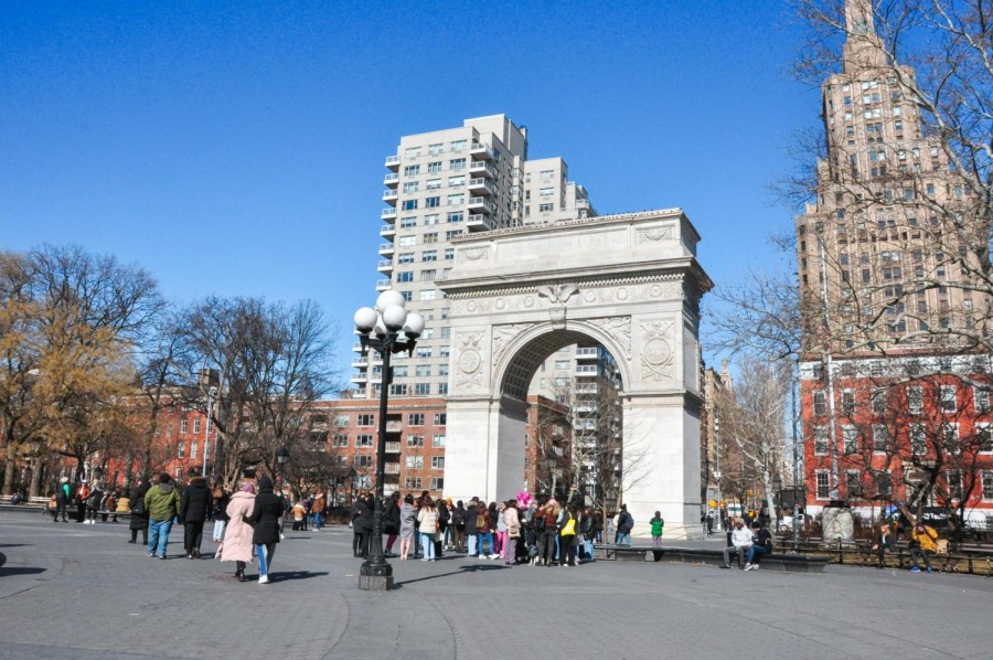 A crowd of people stand and walk around a park surrounded by city buildings and an arch monument under a clear blue sky.