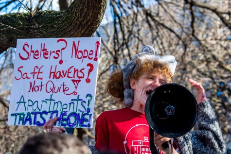 A closeup of a person yelling into a megaphone holding up a sign that states "Shelters? Nope! Safe Havens? Not Quiet? Apartments? That's The One!" in red and blue ink. A crowd of protesters surround the person.