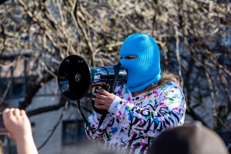 A close-up shot of a person wearing a blue ski mask speaking into a megaphone to a crowd of protesters.