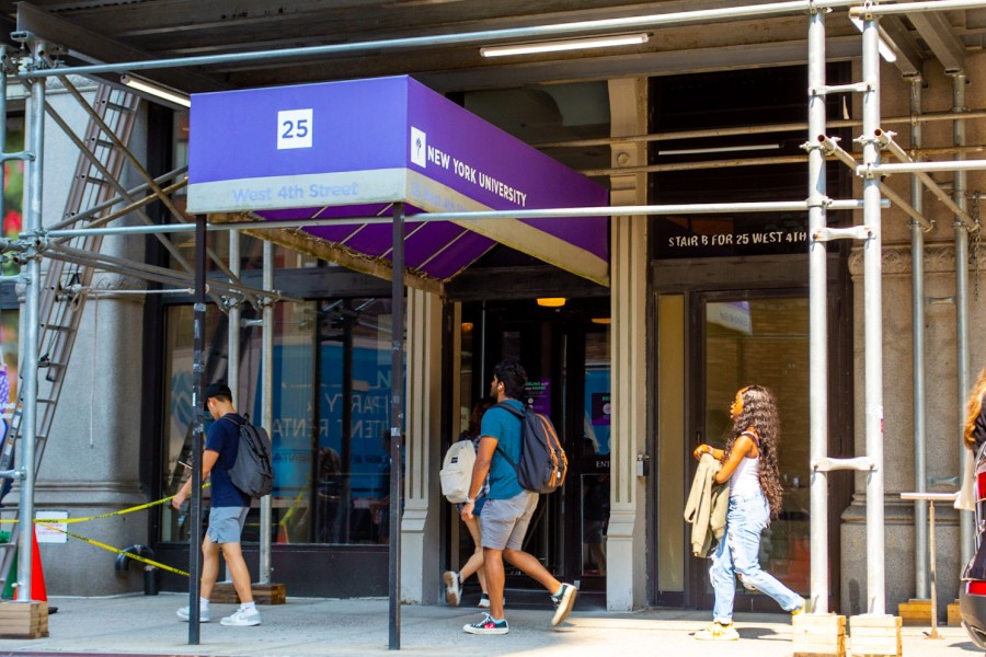 A vertical view of the facade of the 25 West 4th St. NYU building. In front of the entrance door there is a purple roof that on the side says “New York University.” On the front there is a white box with the number “25” inside. Under the “25” it reads “West 4th Street”. There are five people walking past the building on the sidewalk.