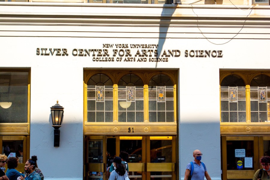 The exterior façade of a college building with golden doors and clear decorated windows as the sun shines on half of the building and people walk on the sidewalk.