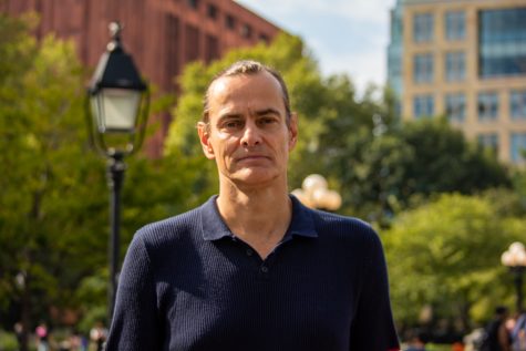 A photo portrait of NYU Professor Ulrich Baer wearing a navy blue polo shirt in front of a lamppost and trees in Washington Square Park.