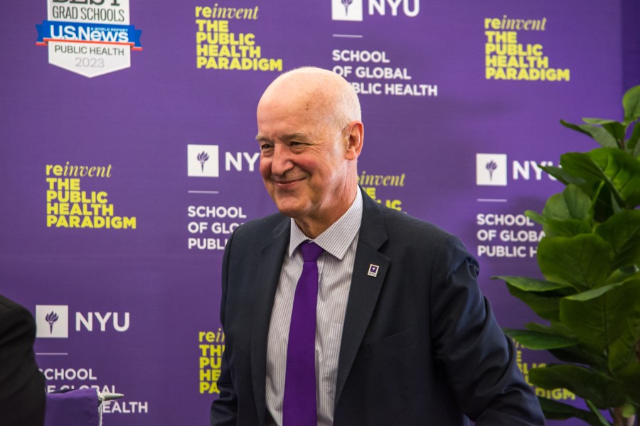 NYU president Andrew Hamilton standing in the NYU School of Global Public Health. Behind Andrew Hamilton, a purple banner hangs with the NYU logo and the words “School of Global Public Health” in white letters. Hamilton is wearing a black blazer, white button-up shirt and bright purple tie.