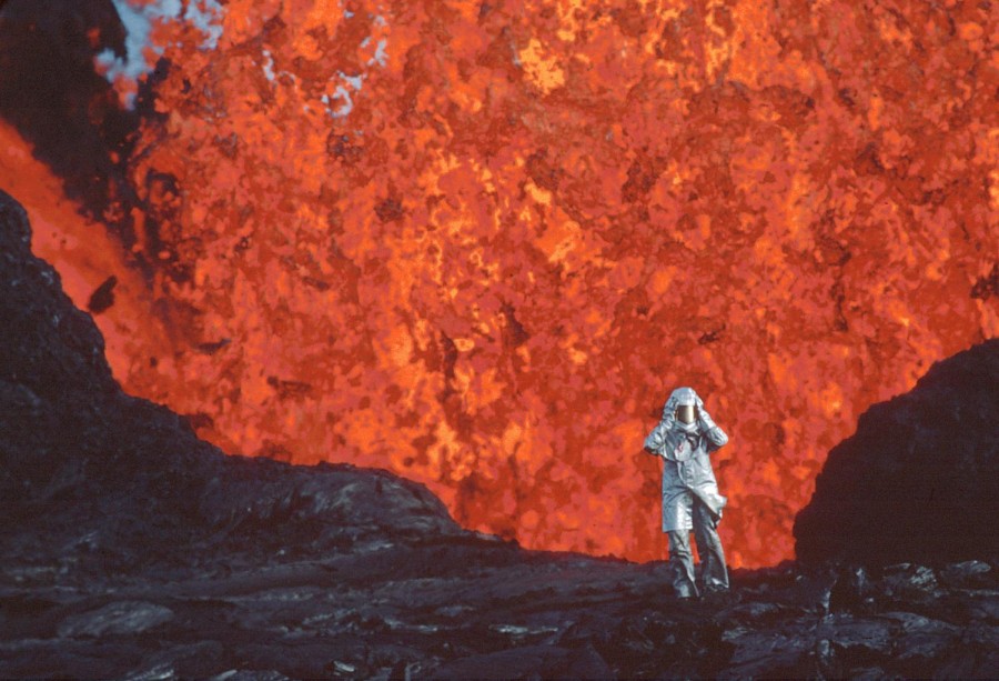 A figure dressed in white fire-resistant suit walking away from an eruption of lava.