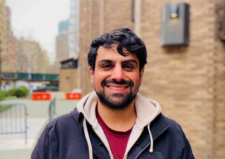 A portrait of Hesam Oveys wearing a black jacket layered over a beige hoodie and a red shirt. He is looking straight at the camera. Behind him is a brick building.