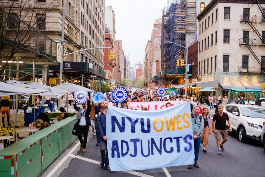 Protesters walk down University Place holding a large sign that says “NYU OWES ADJUNCTS.”