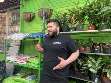 Zack Dahhan stands in front of shelves of potted plants and bags of soil in front of a green brick wall. He appears to be in conversation with someone off camera.