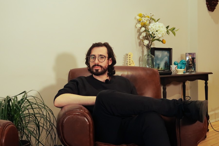 A portrait of Will Wood. A man with brown hair and a beard wearing a black shirt and pants sits on a brown leather chair. Behind him is a table with plants and picture frames.